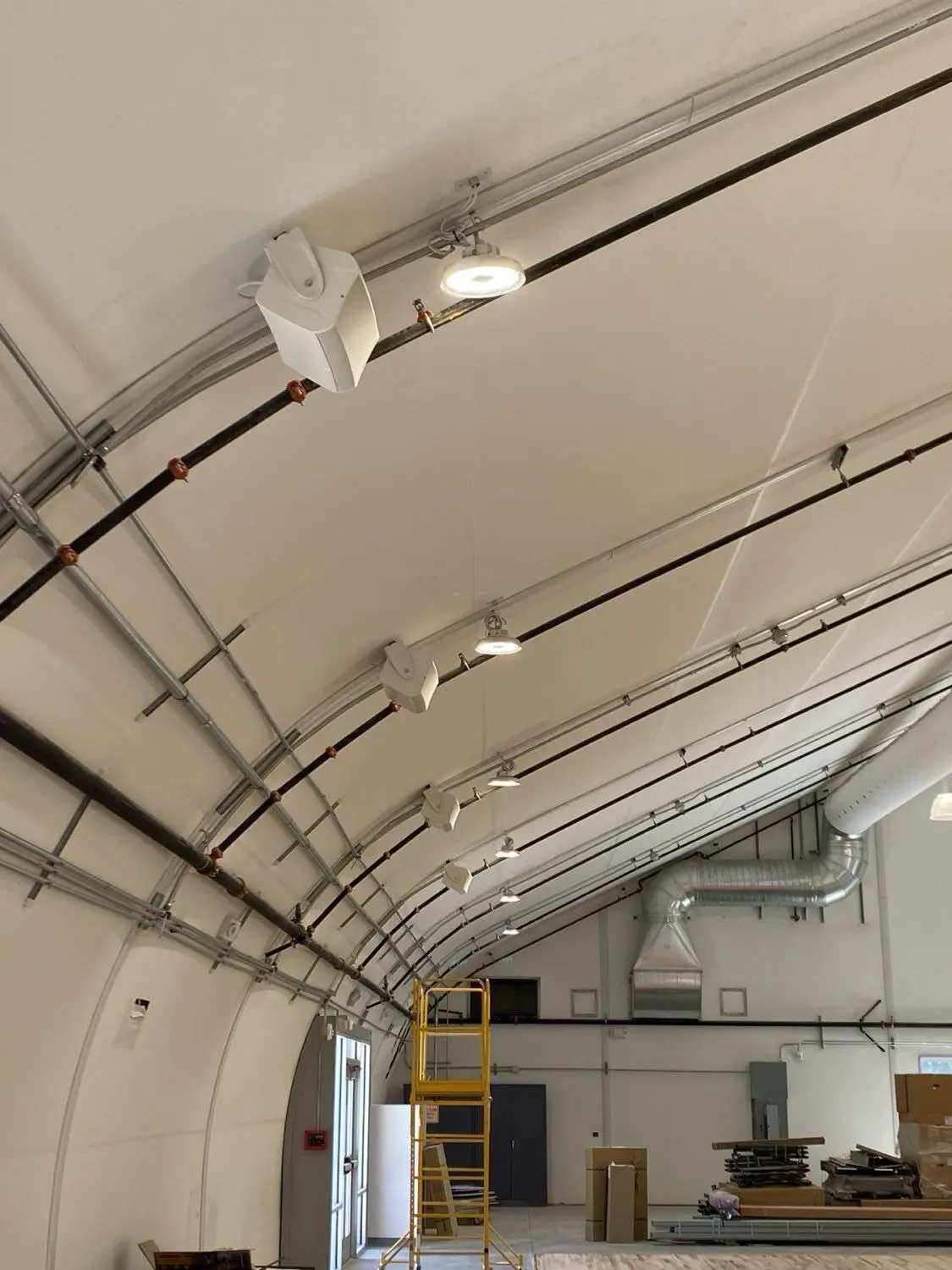 Commercial ceiling-mounted white speakers wired in sequence along the curved structural ribs of an athletic facility