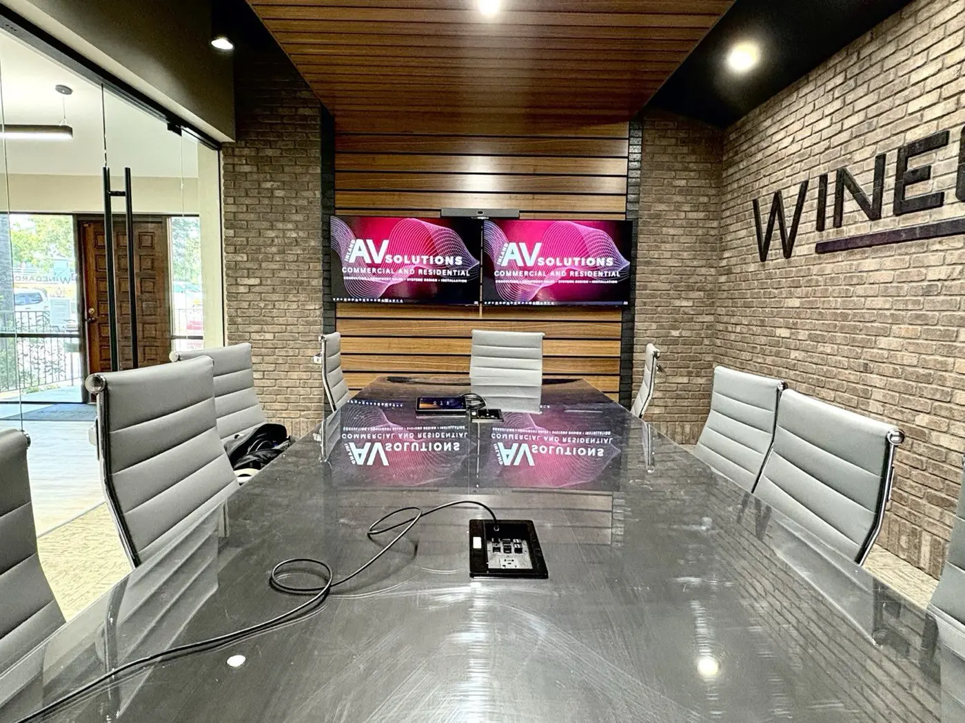 Finished executive boardroom with exposed brick, wood-slat accent wall, glass conference table, and dual displays showing Inland AV Solutions branding