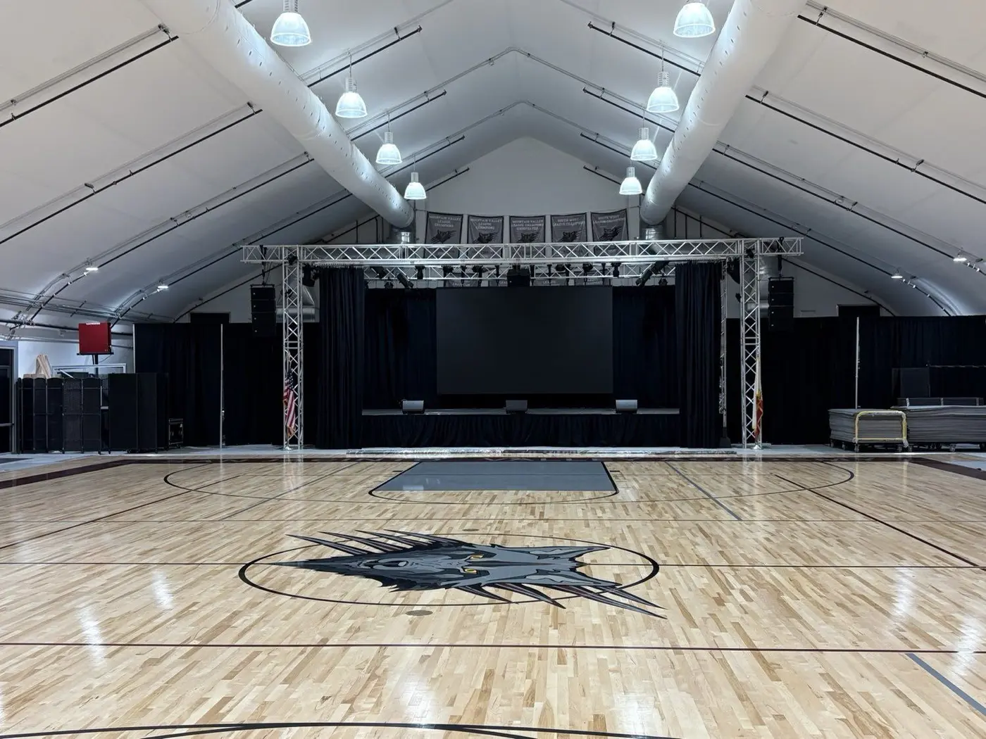 Wide view of the SJVA athletic facility, full basketball court, championship banners, and a truss-mounted stage with projection screen